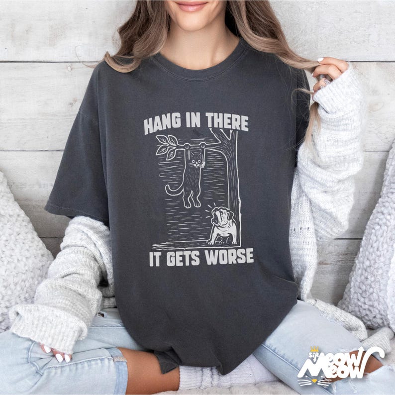 May include: Dark gray t-shirt with a white graphic of a cat hanging from a tree branch, and a dog looking up. The text "HANG IN THERE" is above the cat, and "IT GETS WORSE" is below. The shirt has short sleeves.