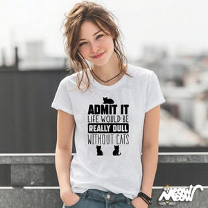 May include: White t-shirt with the text "ADMIT IT LIFE WOULD BE REALLY DULL WITHOUT CATS" in black. The shirt features three black cat silhouettes. The model is wearing blue jeans and a black necklace.
