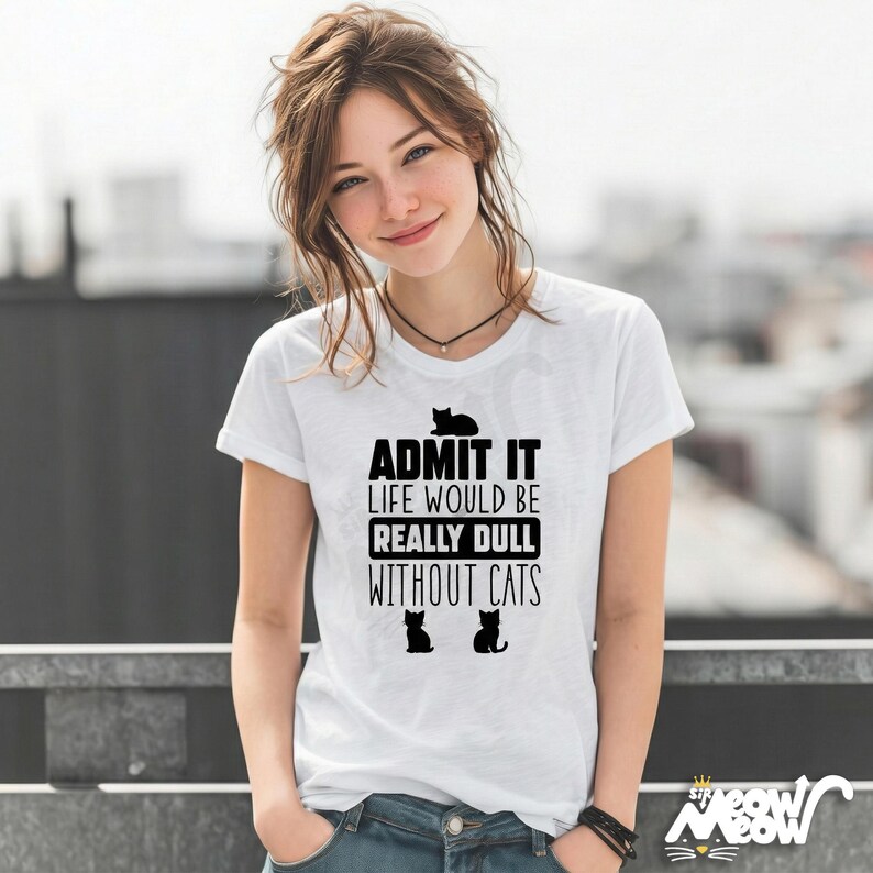 May include: White t-shirt with the text "ADMIT IT LIFE WOULD BE REALLY DULL WITHOUT CATS" in black. The shirt features three black cat silhouettes. The model is wearing blue jeans and a black necklace.
