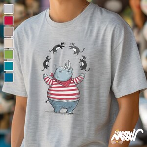 Op de afbeelding: Een grijs T-shirt met een cartoonillustratie van een neushoorn die een rood-wit gestreept shirt en een rode bandana draagt. Vier zwarte katten springen rond de neushoorn.