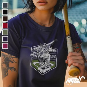 May include: A navy blue t-shirt featuring a graphic of a cat playing baseball. The cat is wearing a baseball uniform and holding a bat. The graphic is surrounded by a green and white baseball field.