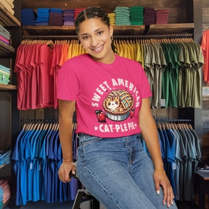 May include: A pink t-shirt with a graphic of a cat inside a pie. The text on the shirt reads "Sweet American *Cat-Ple Pie*"