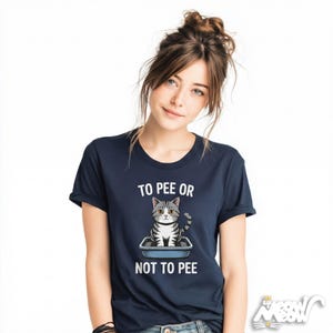 May include: Navy blue t-shirt with a cartoon cat in a litter box and the text "TO PEE OR NOT TO PEE". The cat is grey and white. The shirt has short sleeves and a round neckline.