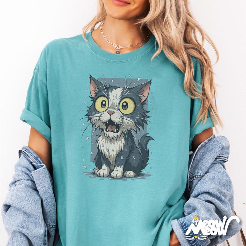 May include: Teal t-shirt with a cartoon cat illustration. The cat has large yellow eyes and an open mouth, looking surprised. It's grey and white, with water droplets around it. The shirt has rolled-up sleeves and the word "Meow" in the bottom right corner.