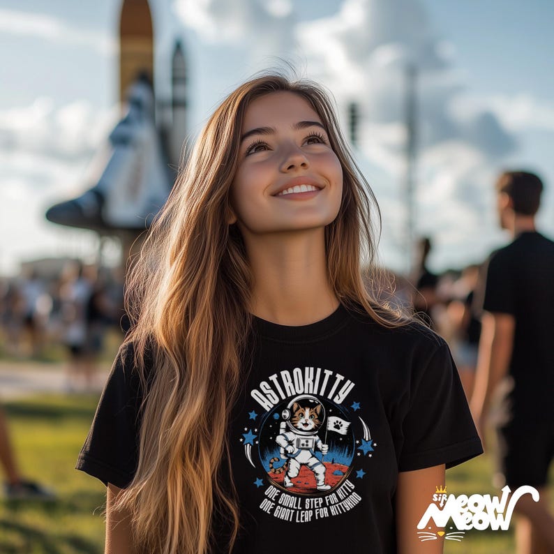 May include: A woman wearing a black t-shirt with a graphic of a cat in an astronaut suit. The graphic says "Astrokitty" and "One small step for kitty, one giant leap for kittkind".