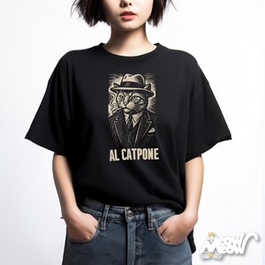 K&ouml;nnte beinhalten: Schwarzes T-Shirt mit einer detaillierten Illustration einer Katze mit Hut und Anzug, die eine Zigarre raucht. Der Text "AL CATPONE" ist unter dem Bild aufgedruckt. Das Shirt ist ein Freizeitartikel.