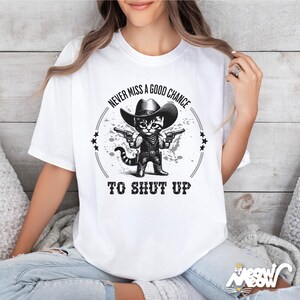 K&ouml;nnte beinhalten: Wei&szlig;es T-Shirt mit einer schwarz-wei&szlig;en Grafik eines Katers mit Cowboyhut und zwei Pistolen. Der Text auf dem Shirt lautet "Never miss a good chance to shut up."
