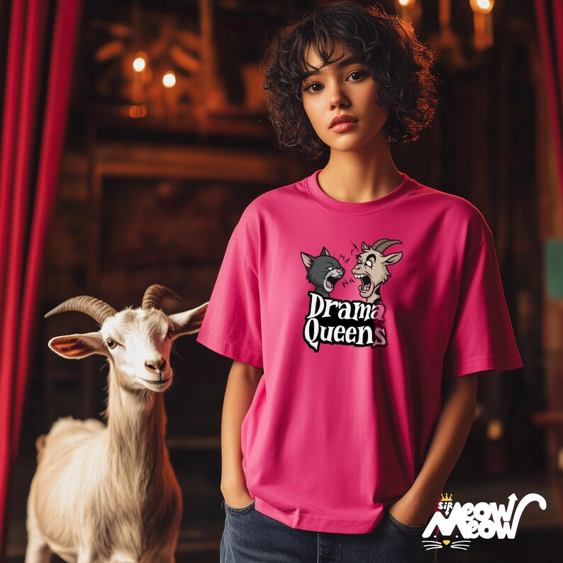 K&ouml;nnte beinhalten: Eine Frau tr&auml;gt ein pinkfarbenes T-Shirt mit einem Aufdruck einer Katze und einer Ziege mit dem Text "Drama Queens". Sie steht vor einer wei&szlig;en Ziege.