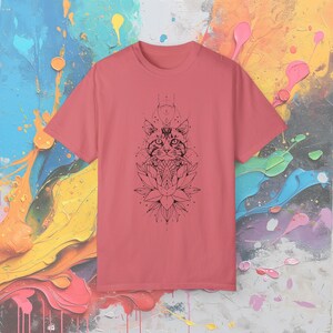 May include: A coral-colored t-shirt featuring a black graphic of a cat's face above a lotus flower. The design has geometric and dot details. The shirt is made of a soft material and has a crew neck.