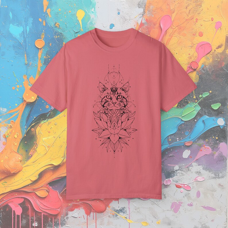 May include: A coral-colored t-shirt featuring a black graphic of a cat's face above a lotus flower. The design has geometric and dot details. The shirt is made of a soft material and has a crew neck.