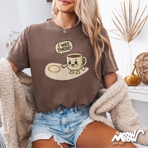 May include: A brown t-shirt with a cartoon coffee cup graphic and the text "I was there!" The coffee cup is holding a finger up to a coffee ring on the ground.
