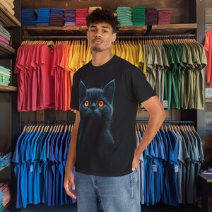 May include: Black t-shirt featuring a detailed illustration of a black cat with bright orange eyes. The cat's fur is depicted with fine detail, creating a realistic appearance. The t-shirt is displayed in a retail setting, surrounded by various colored t-shirts.