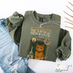 May include: An olive green sweatshirt with the text "FOLLOW YOUR OWN STORY PATH THE CAT THAT DIED & LIVED" and a graphic of an orange cat. The sweatshirt is paired with light blue jeans and a silver necklace.