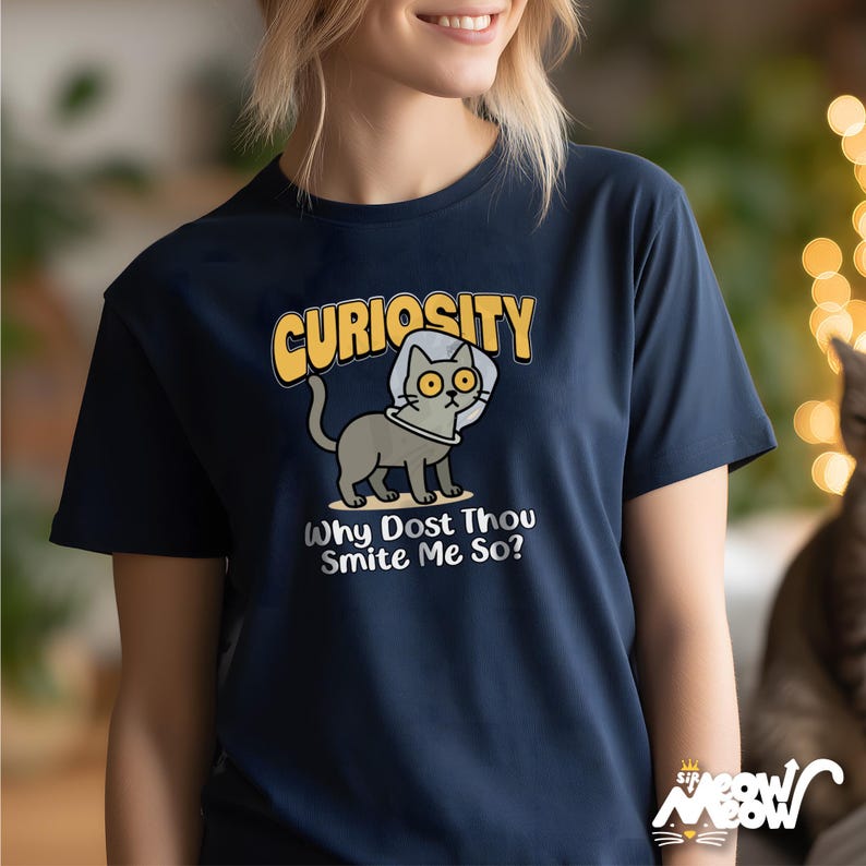 Op de afbeelding: Marineblauw T-shirt met een cartoonkat met een kraag. Het woord "Curiosity" staat boven de kat, met de zin "Why Dost Thou Smite Me So?" eronder. Het shirt heeft een ronde hals.