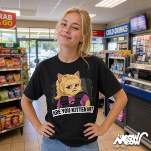 K&ouml;nnte beinhalten: Schwarzes T-Shirt mit einem Cartoon-Katzen-Design, das eine Weste tr&auml;gt und in einem Autositz sitzt, mit dem Text "ARE YOU KITTEN ME?". Das T-Shirt wird von einer Person getragen. Im Hintergrund ist ein Gesch&auml;ft zu sehen.