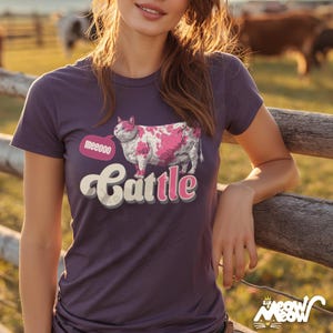 May include: A purple t-shirt featuring a pink and white cow-cat illustration with the word "Cattle" in a playful font. The speech bubble says "meeeoo". The shirt is a crew neck and has short sleeves.