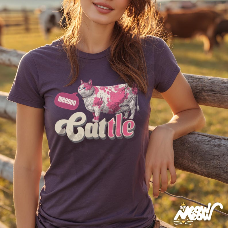 May include: A purple t-shirt featuring a pink and white cow-cat illustration with the word "Cattle" in a playful font. The speech bubble says "meeeoo". The shirt is a crew neck and has short sleeves.