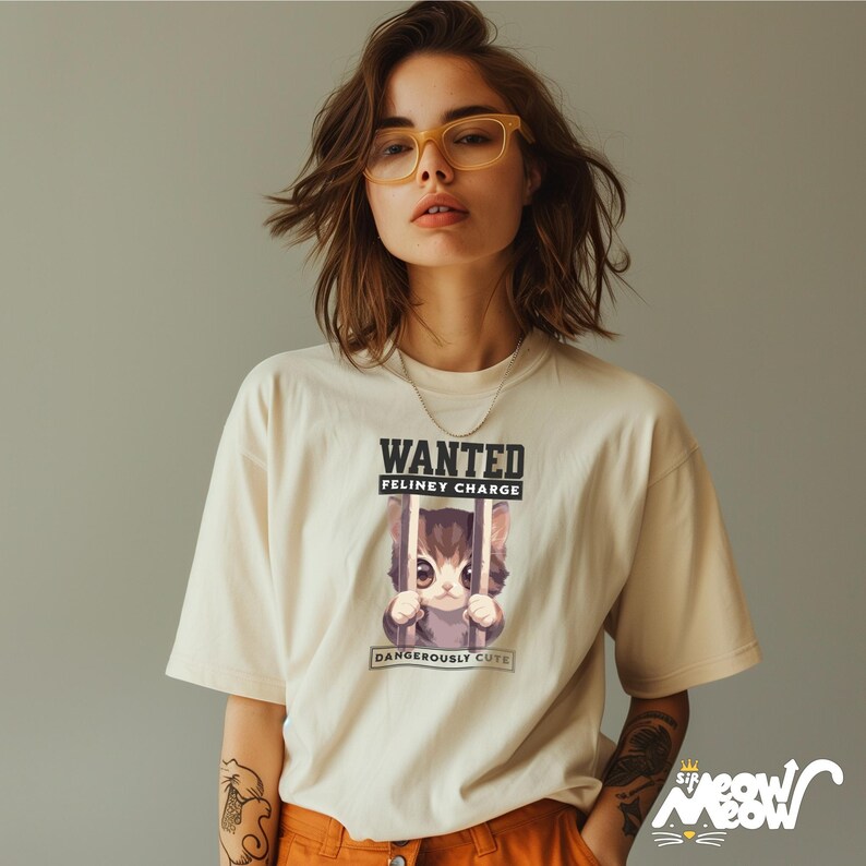May include: A woman wearing a white t-shirt with a graphic of a cat in a jail cell. The graphic says "WANTED FELINEY CHARGE DANGEROUSLY CUTE".