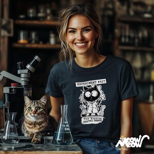 May include: A woman wearing a dark blue t-shirt with a black and white illustration of a cat in a lab coat. The text on the shirt reads "Experiment #427 The Evasive Hairball Remedy".
