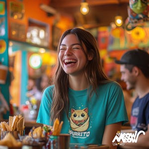 May include: A woman wearing a teal blue t-shirt with a cartoon cat holding a burrito printed on the front. The text "purrito" is printed below the cat. The woman is laughing and sitting at a table with a plate of chips and salsa in front of her.