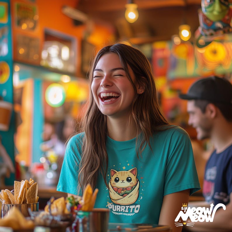 May include: A woman wearing a teal blue t-shirt with a cartoon cat holding a burrito printed on the front. The text "purrito" is printed below the cat. The woman is laughing and sitting at a table with a plate of chips and salsa in front of her.