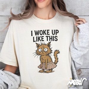May include: A white t-shirt with the text "I woke up like this" and a cartoon illustration of a cat wearing a pink tutu.