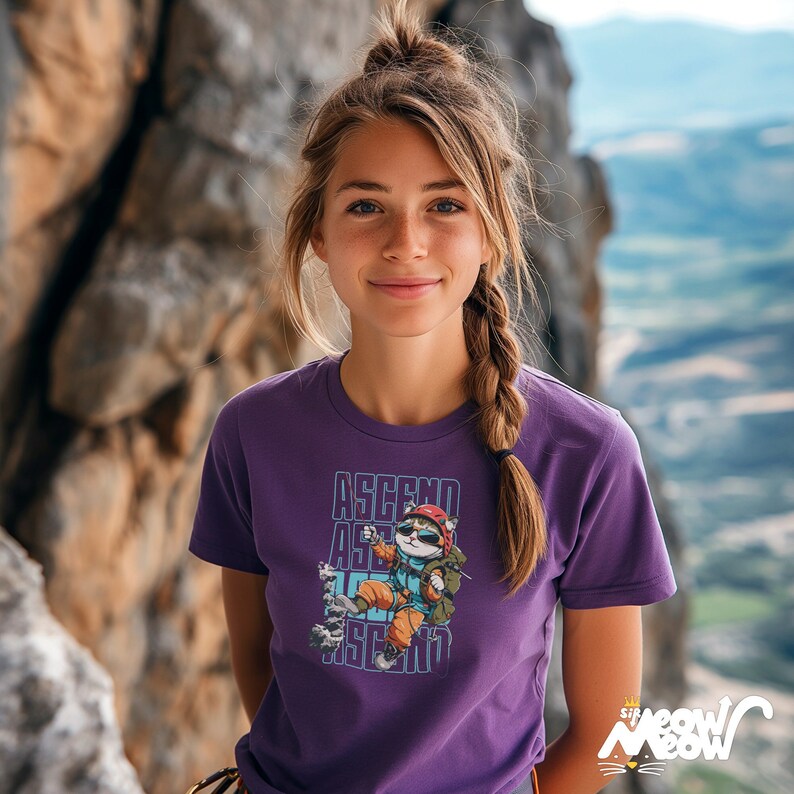 Pu&ograve; includere: Una donna indossa una maglietta viola con una grafica di un gatto che scala una roccia e il testo "ASCEND ASCEND ASCEND".