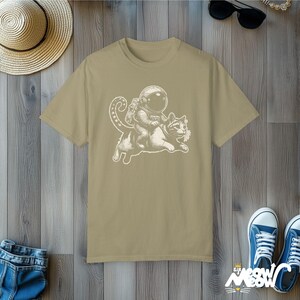 Khaki green Comfort Colors 1717 unisex T-shirt with a humorous white graphic of an astronaut riding atop a playful cat, combining outer space and cat lover themes.