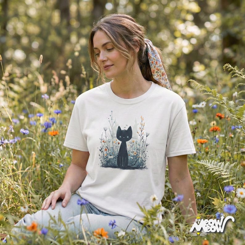 May include: Cream-colored t-shirt featuring a black cat illustration amidst wildflowers. The cat is depicted in a simple, stylized manner. The shirt is worn by a person sitting in a field of colorful flowers.