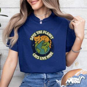 以下が含まれることがあります： 地球で眠る猫のグラフィックが描かれた青いTシャツ。シャツのテキストは「Save the Planet Cats Live Here.」です。