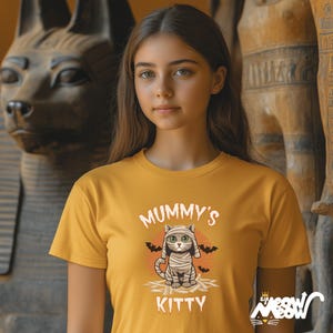 Pode incluir: Camiseta amarelo mostarda com o texto "Mummy's Kitty" e um gr&aacute;fico de um gato m&uacute;mia. O gato est&aacute; enrolado em bandagens e tem olhos verdes. Dois morcegos pretos est&atilde;o em ambos os lados do gato. A camiseta &eacute; usada por uma pessoa.