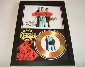 May include: A framed gold disc award for Depeche Mode. The disc is surrounded by a black mount with a black and red design. The disc features a photo of the band members. The band's name is written in yellow on the mount.