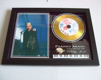 May include: A framed gold disc award featuring a black and white photo of Billy Joel performing on stage. The disc is titled "Piano Man: The Music of Billy Joel" and includes a small image of Billy Joel playing the piano.