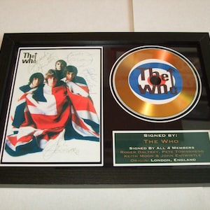 May include: Framed memorabilia featuring The Who. Includes a photo of the band draped in a Union Jack, a CD with the band's logo, and a plaque stating "Signed by The Who". The frame is black.