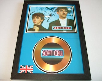 May include: Framed Soft Cell memorabilia. The image features a photo of the band members with signatures, a gold-coloured disc, and the band's logo. The frame is black, with a blue background. A small Union Jack flag is in the lower left corner.