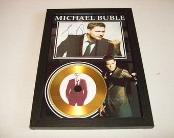 May include: A framed gold record award with a black frame and a photo of Michael Bublé. The record is gold with a white label and a photo of Michael Bublé in a suit. The photo is signed by Michael Bublé.