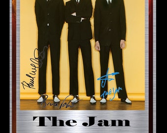 May include: A framed photograph of The Jam band members, Paul Weller, Bruce Foxton, and Rick Buckler, wearing black suits and white shoes. The photograph is signed by each band member. The frame is silver with a black mount and a red and gold border. The text "The Jam Limited Edition Signed Display" is printed below the photograph.
