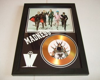 May include: A framed gold disc award for Madness, a British ska band. The disc is surrounded by a black frame with a white logo and the band's name in white letters. The disc features a photo of the band members. The frame also includes a smaller photo of the band members with their signatures.