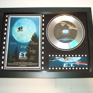 Könnte beinhalten: Gerahmte E.T.-Filmerinnerungsstücke. Der Rahmen enthält ein Filmplakat, eine CD und einen Filmstreifen. Das Plakat zeigt E.T. und Elliott vor dem Mond. Die CD hat den Filmtitel und das Bild. Der Rahmen ist schwarz.
