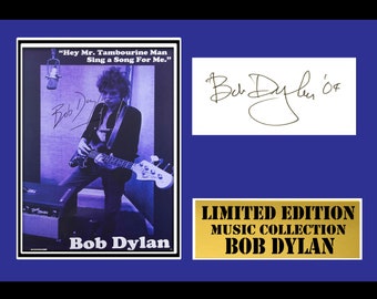 May include: A black and white photograph of Bob Dylan playing a bass guitar in a recording studio. The photo is framed in a blue mount with a gold plaque that reads "Limited Edition Music Collection Bob Dylan". The photo is signed by Bob Dylan.