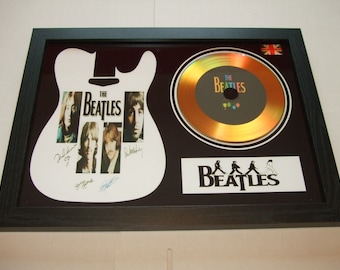 May include: Framed Beatles memorabilia. It includes a white guitar-shaped image with band member photos and signatures, a gold CD, and the band's logo. The black frame also features a small Union Jack flag in the top right corner.