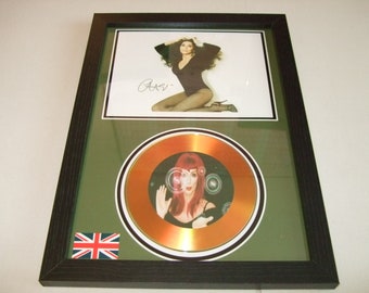 May include: A framed gold disc featuring a picture of a woman with red hair. The disc is surrounded by a black border and a green mount. The frame is black with a Union Jack flag in the bottom left corner. The image includes the signature "Cher".