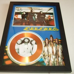 Puede incluir: Un disco de oro enmarcado con una foto de los miembros de la banda Bee Gees. El disco está rodeado de un fondo azul con el nombre de la banda en letras doradas. El marco también incluye una foto más pequeña de los miembros de la banda con sus autógrafos.