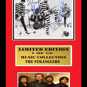 Puede incluir: Una fotografía en blanco y negro de los miembros de la banda The Stranglers con uniformes de prisión, sosteniendo instrumentos musicales. La foto está firmada por cada miembro de la banda. La foto está enmarcada con una placa dorada que dice "Edición limitada 1 de 50 Colección musical The Stranglers". Debajo de la foto hay una foto más reciente de los miembros de la banda.