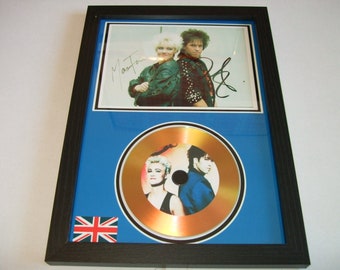 May include: A framed gold disc featuring a photo of a band, signed by the band members. The photo shows two people, a woman with blonde hair and a man with dark hair. The disc is framed with a blue mount and a black frame. The frame also includes a small Union Jack flag.