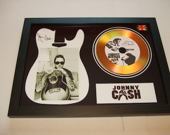 May include: A framed gold disc featuring a black and white photo of Johnny Cash. The disc is shaped like a guitar and has the text "Johnny Cash" on it. The disc is signed by Johnny Cash.