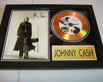 May include: Framed Johnny Cash memorabilia. The frame contains a black and white photo of Johnny Cash, a gold-coloured CD with Cash's image, and a gold plaque with "JOHNNY CASH" in black text. The frame is black.