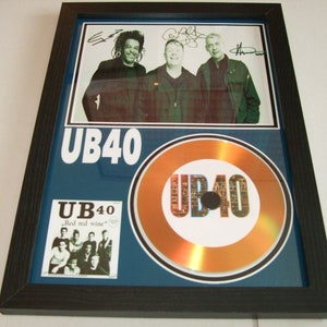 Puede incluir: Un disco de oro enmarcado para la banda UB40. El disco está rodeado por un borde azul con el nombre de la banda en letras blancas. Una foto más pequeña de la banda está en la esquina superior izquierda con sus firmas. Una foto más pequeña de la portada del álbum de la banda está en la esquina inferior izquierda.
