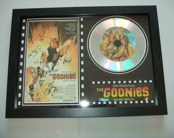 May include: A framed movie poster and CD featuring the Goonies film. The poster is signed by the cast and includes the title "The Goonies" and the text "A Richard Donner Film". The CD is a silver disc with a picture of the Goonies cast on it.