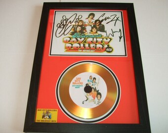 May include: A framed gold disc featuring a picture of the Bay City Rollers. The disc is signed by the band members. The frame is black with a red mount. The text on the disc reads "Bay City Rollers Saturday Night". The text on the mount reads "Bay City Rollers The Anthology 4CD".
